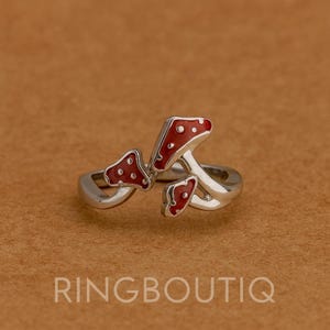 Può includere: Anello argentato con tre ciondoli a forma di fungo rosso con pois bianchi. L'anello ha un design avvolgente. Lo sfondo è marrone e il testo "RINGBOUTIQ" è visibile in basso.