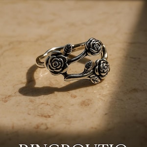 Könnte beinhalten: Silberner Ring mit drei Rosenmotiven, jedes mit einem Blatt, auf einem astförmigen Band. Der Ring liegt auf einer hellbraunen Oberfläche. RINGBOUTIq ist in weißer Schrift auf der Oberfläche geschrieben.