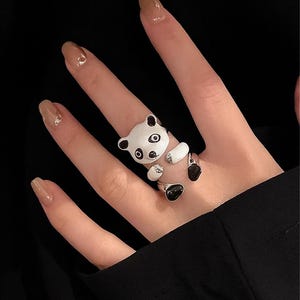 Può includere: Anello color argento con un design a forma di panda bianco e nero. Il panda ha orecchie, occhi e piedi neri, con un corpo bianco. L'anello è su una mano con unghie curate. La parola "RINGBOUTIQ" è in alto.