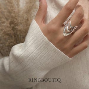 May include: A silver ring with a layered, wave-like design is shown on a hand. The ring has a polished finish and a modern aesthetic. The word "RINGBOUTIQ" is printed at the bottom of the image.