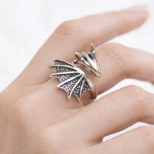 May include: A silver dragon ring with gold-colored accents on the horns. The dragon's wings are detailed with a textured pattern. The ring is wrapped around a finger, showcasing the intricate design of the fantasy-themed jewelry.