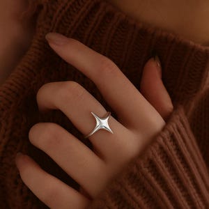 Könnte beinhalten: Ein silberner Ring mit einem Sternenmuster. Der Ring, der an einem Finger getragen wird, hat einen Stern mit vier Spitzen. Der glänzende Metallring ist vor einem braunen Hintergrund platziert.