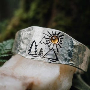 May include: A wide silver cuff ring with a hammered texture featuring an engraved landscape design of two pine trees and mountains. A small, round, faceted amber-colored gemstone is set into the center of an engraved sun above the mountains.