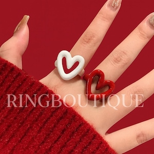 以下が含まれることがあります： 赤色の背景に、白いハートと赤いハートの2つの指輪が写っています。指輪は人の指にあり、白いハートは赤い内側があり、赤いハートは単色です。「RINGBOUTIQUE」の文字が見えます。