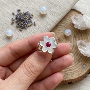 Könnte beinhalten: Ein Silberring mit einer blumenförmigen Perlmutt-Einlage und einem kleinen, runden, facettierten rosa Edelstein in der Mitte. Der Ring besteht aus poliertem Silber und die Blütenblätter haben eine glatte, schimmernde weiße Oberfläche.