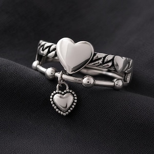 Puede incluir: Un anillo de tono plateado con diseño de doble banda. La banda superior muestra una forma de corazón lisa, mientras que la banda inferior consiste en una serie de pequeñas esferas. Un pequeño dije de corazón con cuentas cuelga de la banda inferior.