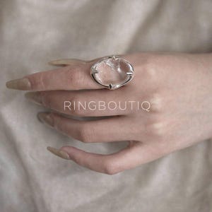 May include: A silver-toned ring with a clear, oval-shaped stone. The ring has a unique design with pointed accents. The text "RINGBOUTIQ" is visible below the ring.