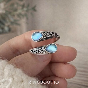 Puede incluir: Dos anillos de plata con una piedra azul iridiscente en forma de lágrima. Los anillos tienen un diseño ornamental y arremolinado alrededor de la piedra. El texto "RINGBOUTIQ" está en la parte inferior de la imagen.