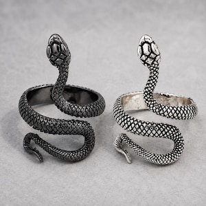 May include: Two wrap-around rings shaped like snakes. One ring is finished in a dark, matte black metal, while the other is finished in a bright, polished silver tone. Both rings feature detailed, textured scale patterns and raised snake heads.
