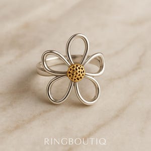 May include: A silver-toned ring with a daisy flower design. The flower has five curved petals and a gold-colored center. The ring is set against a neutral background. The text "RINGBOUTIQ" is at the bottom.