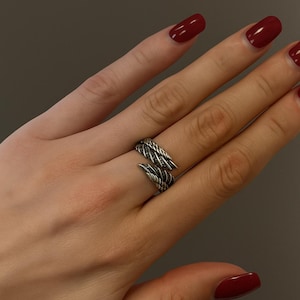 Op de afbeelding: Een zilverkleurige ring met een getextureerd, gevlochten ontwerp, die zich om de vinger wikkelt. De ring heeft een gespleten ontwerp met puntige uiteinden. De afbeelding toont ook een hand met rode nagellak en de tekst "RINGBOUTIQ".