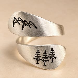 May include: A silver-toned bypass ring with a brushed matte finish. The upper end features an engraved black mountain range silhouette, while the lower end displays an engraved black forest scene with three evergreen pine trees.