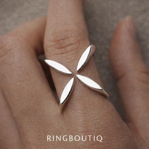 May include: A silver ring with a four-petal design. The petals are elongated and curved, forming a symmetrical shape. The ring is worn on a finger, with the brand name "RINGBOUTIQ" visible at the bottom.