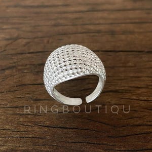 Op de afbeelding: Een zilverkleurige ring met een getextureerd, gewelfd ontwerp. De ring heeft een reeks kleine gaatjes over het oppervlak. De tekst "RINGBOUTIQU" is zichtbaar onderaan de afbeelding.