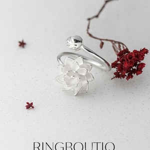 May include: A silver-colored ring with a flower design. The ring features a detailed, open flower and a bud. The word "RINGBOUTIQ" is printed in black. Red accents and a branch are in the background.