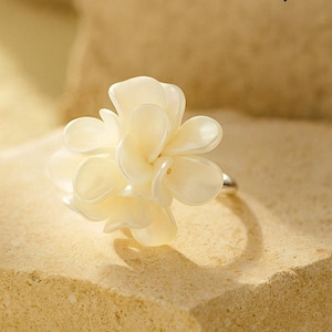 May include: A cream-coloured flower ring with a silver band sits on a textured, light brown surface. The ring's floral design is the focal point, with the word "RINGBOUTIQ" in black letters at the top.