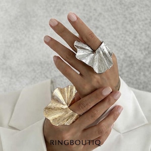 Puede incluir: Dos anillos grandes y llamativos con un diseño texturizado en forma de abanico. Un anillo es plateado, el otro es dorado. Los anillos se llevan en los dedos, con el nombre de la marca "RINGBOUTIQ" visible en la parte inferior de la imagen. El fondo es gris suave.