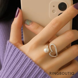 May include: A silver ring featuring a heart shape and a pearl. The ring is worn on a finger, with a purple sweater visible. The image includes the text "RINGBOUTIQ" at the bottom.