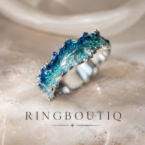Può includere: Un anello in argento con un bordo irregolare e testurizzato, caratterizzato da una vibrante sfumatura di smalto blu e turchese sulla superficie. La parola RINGBOUTIQ è stampata in lettere maiuscole nere sotto l'anello, accompagnata da una piccola stella a quattro punte.