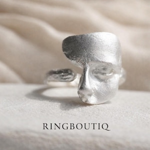 May include: A silver ring featuring a sculpted face design. The ring has a textured finish and the word "RINGBOUTIQ" is printed at the bottom. The face has detailed features, including an eye, nose, and mouth.