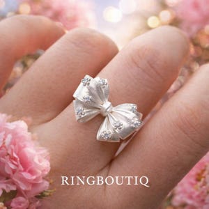 May include: A silver bow ring with small, sparkling accents. The ring is worn on a finger, with the bow design prominently displayed. The word "RINGBOUTIQ" is printed at the bottom of the image.