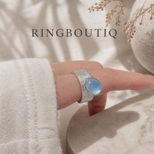 May include: A silver ring with a textured band and a large, oval, iridescent stone. The ring is worn on a finger, and the word "RINGBOUTIQ" is displayed in the background.