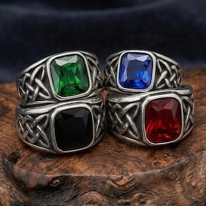 Puede incluir: Cuatro anillos de metal plateado con intrincados patrones trenzados en las bandas. Cada anillo tiene una gran piedra preciosa rectangular facetada de colores distintos: verde esmeralda, azul real, negro profundo y rojo rubí.