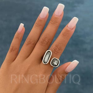 Może przedstawiać: Srebrny pierścionek z dwoma szarymi kamieniami, jednym prostokątnym i jednym kwadratowym, jest prezentowany na dłoni z zadbanymi paznokciami. Słowo "RINGBOUTIQ" jest widoczne w lewym dolnym rogu.