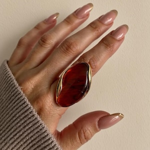 Pode incluir: Um anel oval grande com uma pedra polida de cor âmbar profundo, engastada numa moldura de metal dourado liso. O anel tem um design proeminente e alongado que cobre uma parte significativa do dedo, exibindo um tom castanho-avermelhado rico e translúcido.