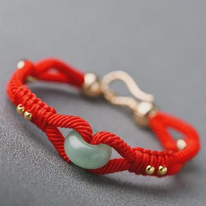 Peut inclure: Un bracelet rouge tressé avec une perle de jade vert clair au centre. Le bracelet comporte des accents dorés et un fermoir. Le fond est gris neutre.