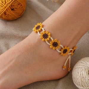 Könnte beinhalten: Gehäkeltes Sonnenblumen-Fußkettchen mit gelben und braunen Blumendetails, verbunden durch eine weiße Kordel. Das Fußkettchen wird an einem Knöchel gezeigt, mit einem Knäuel gelber Wolle und einem Häkelhaken im Hintergrund.