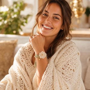 May include: A cream-colored crochet bracelet with a large flower design and gold accents. The bracelet features a delicate, open-weave pattern and is adorned with small beads and a tassel. The image also shows a cream-colored crochet shawl.