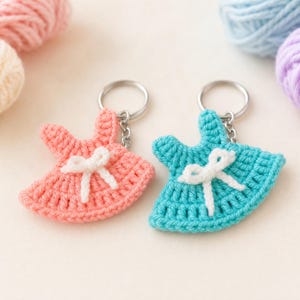 Peut inclure: Deux porte-clés en crochet en forme de petites robes, l'une rose corail et l'autre bleu turquoise. Chaque robe présente un nœud en crochet blanc au centre et est fixée à un anneau fendu en métal argenté par une courte chaîne.