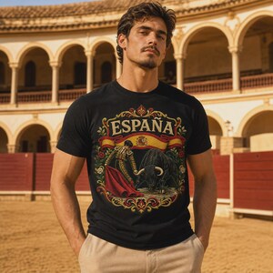 May include: A black short-sleeved t-shirt featuring a graphic print of a bullfighter with a red cape and a bull. The design includes the text ESPAÑA, a Spanish flag banner, and ornate gold and red floral borders surrounding the central illustration.