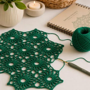 May include: A close-up of a handcrafted emerald green crochet doily with a floral pattern. A ball of matching yarn, a crochet hook, and a notebook with the text "Emerald Radiance Crochet Pattern" are also visible.