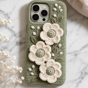 Peut inclure: Une coque de téléphone en crochet vert sauge ornée de trois fleurs crochetées de couleur crème. La coque présente un motif de point texturé et de délicates feuilles et tiges vertes. Le texte "Crée par HERBAAAL" est visible en haut.