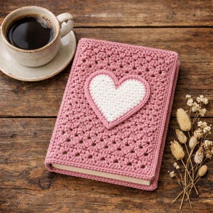 Peut inclure: Une couverture de livre rose crochetée avec un cœur blanc appliqué. La couverture est à côté d'une tasse de café et d'un petit bouquet de fleurs séchées. La couverture du livre est sur une surface en bois.