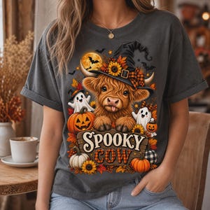 Peut inclure: Un t-shirt gris anthracite avec une illustration d'une vache highland poilue portant un chapeau de sorcière, entourée de citrouilles, de tournesols et de deux fantômes. Le texte "SPOOKY COW" est imprimé sur un panneau en bois.
