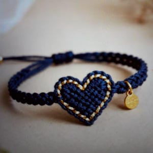 May include: A navy blue macrame bracelet featuring a crocheted heart with gold-colored trim. The heart is the focal point, with a small gold charm dangling from the bracelet. The bracelet is adjustable.