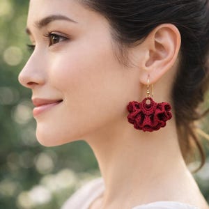 Peut inclure: Gros plan d'une femme portant des boucles d'oreilles en crochet bordeaux. Les boucles d'oreilles ont un design à volants en forme de fleur et sont suspendues à des crochets dorés. L'arrière-plan est flou, avec des tons verts et blancs doux.