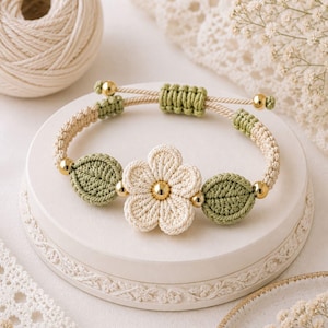 May include: A handmade crochet bracelet featuring a cream-colored flower and two green leaves. The bracelet has a beige cord with green accents and gold-colored beads. The flower has a gold center. The bracelet is displayed on a white, decorative surface.