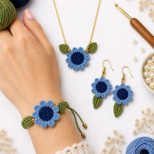 Peut inclure: Un ensemble de bijoux crochetés à la main avec un motif de fleur bleue et de feuilles vertes. L'ensemble comprend un collier, des boucles d'oreilles et un bracelet. Le collier et les boucles d'oreilles ont des chaînes et des crochets dorés. Le bracelet est réglable.