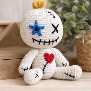 May include: A white crochet doll with a round head and body. It features a yellow tuft on top, a blue star over one eye, a black stitched mouth, and a red heart patch on the chest. Black thread details resemble stitches across the limbs and head.