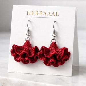 Peut inclure: Une paire de boucles d'oreilles rouges au crochet avec un design à volants. Les boucles d'oreilles sont présentées sur une carte blanche avec le texte doré "HERBAAAAL". Les boucles d'oreilles ont des crochets argentés.