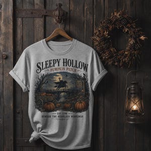 Peut inclure: Un t-shirt gris clair à manches courtes avec une impression graphique représentant le Cavalier sans tête sur un cheval, un champ de citrouilles et un pont. Le texte indique : Sleepy Hollow Pumpkin Patch, Est. 1790, Beware the Headless Horseman.