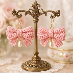 Peut inclure: Une paire de boucles d'oreilles en forme de nœud rose crocheté, avec des crochets dorés et une perle pendante. Les boucles d'oreilles sont présentées sur un présentoir doré décoratif, sur un fond flou.
