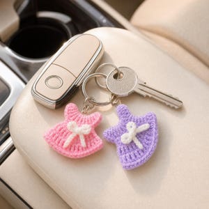 Peut inclure: Un porte-clés en métal avec deux petites breloques en forme de robe crochetée, l'une rose et l'autre lavande, chacune ornée d'un nœud blanc crocheté au centre. Le porte-clés est attaché à une clé en métal argenté et un boîtier de clé beige.