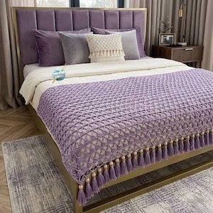 Könnte beinhalten: Ein Bett mit einem lilafarbenen Samt-Kopfteil und einem goldenen Rahmen. Das Bett ist mit einer weißen Bettdecke, grauen und lila Kissen und einer lavendelfarbenen Häkeldecke mit Quasten ausgestattet. Ein kleiner Teppich ist am Fußende des Bettes zu sehen.