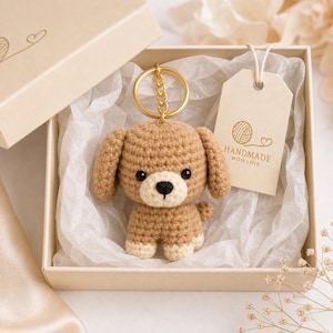 Peut inclure: Un porte-clés chien crocheté beige et crème avec des yeux et un nez noirs, fixé à un anneau en métal doré. Le porte-clés est présenté dans une boîte cadeau beige doublée de papier de soie blanc, avec une étiquette marquée HANDMADE WITH LOVE.