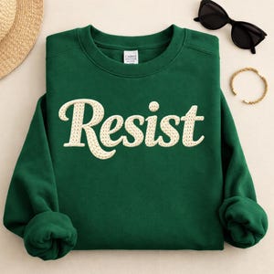 Może przedstawiać: Ciemnozielona bluza z napisem "Resist" wyszytym kremowymi, teksturowanymi literami. Bluza jest złożona, z kapeluszem ze słomy, czarnymi okularami przeciwsłonecznymi i złotą bransoletką w tle.